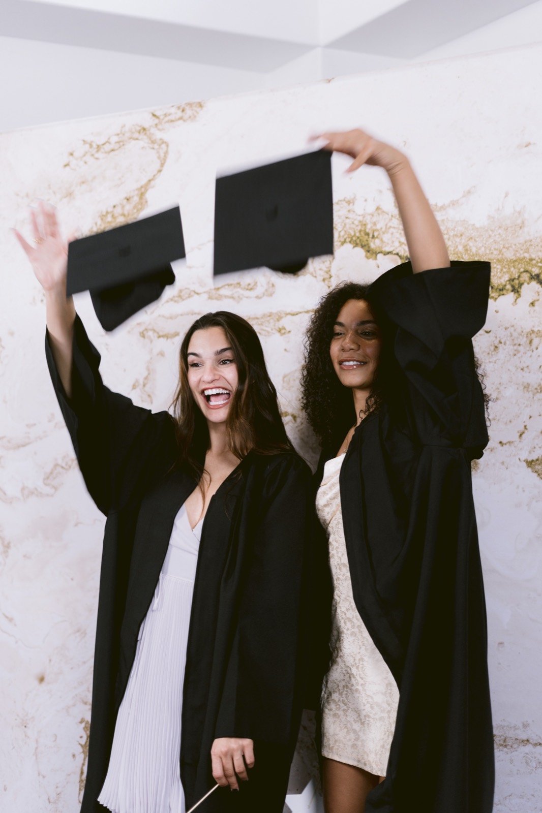 Two graduates in black gowns tossing their caps in front of a cream and gold marble backdrop