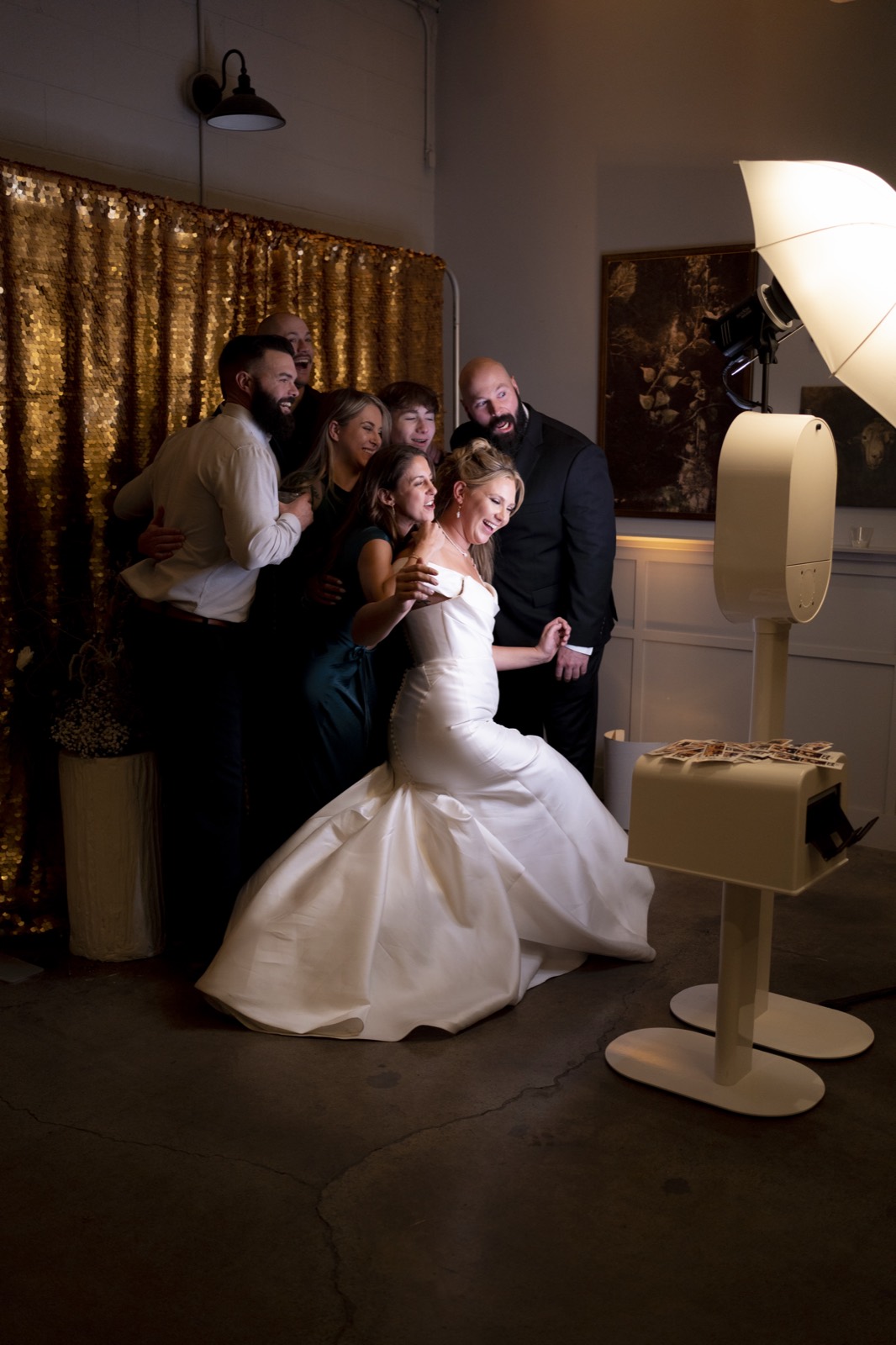 A bride, groom and wedding party crowding into the photo booth in front of a gold sequin backdrop